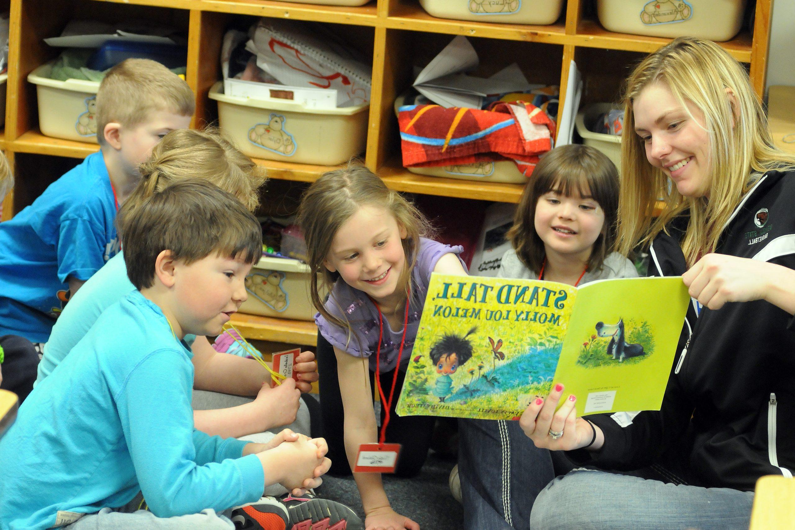 一位老师正在给一群学生朗读一本名叫《昂首挺立》的书，作者是Molly Lou Melon